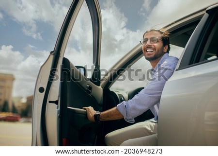 Positive young middle-eastern man going out the car, low angle photo, copy space. Happy arab guy open automobile door and smiling, getting in. Auto test-drive, leasing, buying, renting concept Royalty-Free Stock Photo #2036948273
