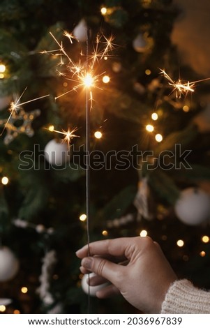 Hand holding  burning sparkler on background of christmas tree  lights in festive evening room. Happy New Year! Firework bengal glowing in woman hand. Space for text. Atmospheric moment