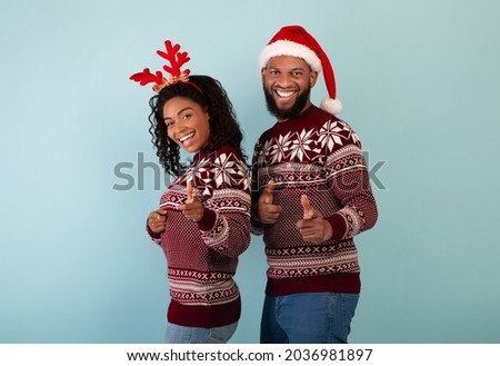 Hey, you. Portrait of happy black couple in Christmas sweaters pointing at camera over blue background. Excited woman wearing deer horns and man in santa hat