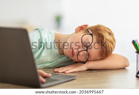 Tired kid fell asleep next to laptop while were doing homework for school, closeup portrait. Exhausted redhead boy with glasses sleeping on desk in front of computer, home interior