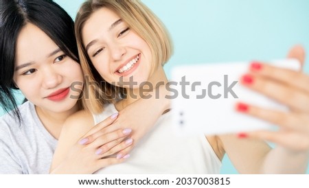 Ethnic diversity. Happy friends. Mobile selfie. Enjoying meeting. Pretty smiling asian caucasian women making photo on smartphone isolated blue.