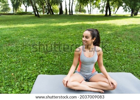 cheerful young woman sitting in lotus pose on yoga mat in park