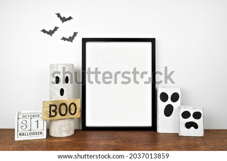 Halloween mock up. Black frame on a wood shelf with rustic wood ghost decor and calendar. Portrait frame against a white wall with bats. Copy space.