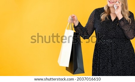 Black Friday. Shopping sale. Large clothing discount. Body positive. Unrecognizable overweight obese woman with bags speaking on phone isolated on bright yellow empty space background.