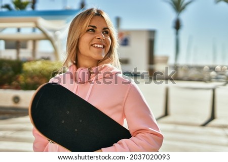 Young blonde skater girl smiling happy holding skate at the city.