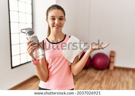 Young brunette teenager wearing sportswear holding water bottle smiling cheerful presenting and pointing with palm of hand looking at the camera. 