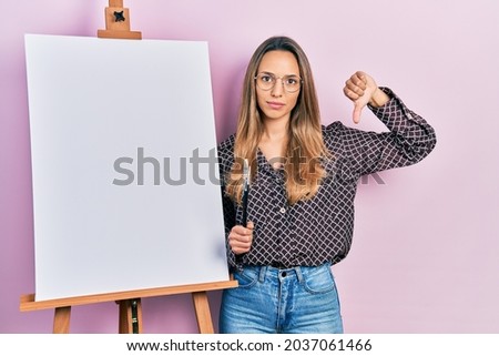 Beautiful hispanic woman standing by painter easel stand holding brushes with angry face, negative sign showing dislike with thumbs down, rejection concept 