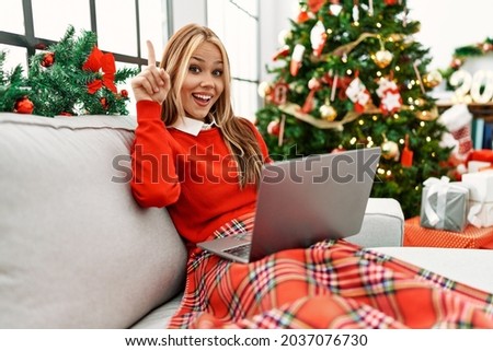 Young caucasian girl using laptop sitting on the sofa by christmas tree pointing finger up with successful idea. exited and happy. number one. 
