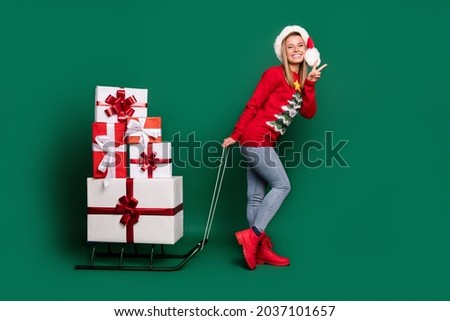 Full length profile photo of funky santa young lady take presents show v-sign wear sweater jeans boots isolated on green color background