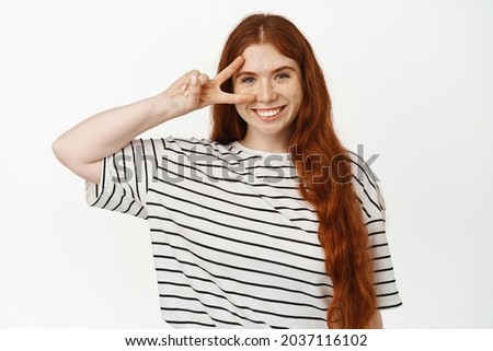 Happy and positive redhead girl showing peace sign near clean no makeup skin, v-sign over eye, standing cheerful in t-shirt against white studio background.