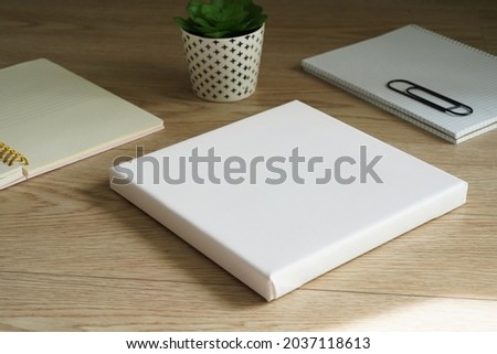 White empty square canvas for painting lies on a wooden table next to two notes and a small flower. Mockup canvas (982)