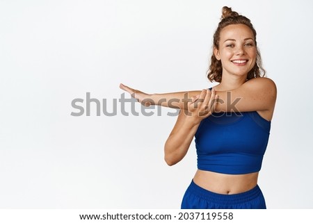 Smiling sportswoman stretching arm and looking enthusiastic. Fitness girl shows workout exercises in blue activewear, stands isolated against white background.