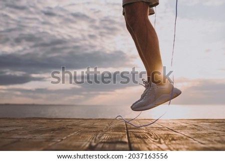 Cropped up photo shot young sporty fit sportsman man in sports clothes shorts warm up training jump on skipping rope at sunrise sun dawn over sea beach outdoor on pier seaside in summer day morning.