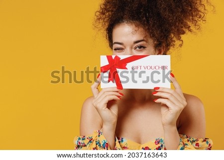 Young happy smiling fun cheerful woman 20s with culry hair in casual clothes hold cover face with gift certificate coupon voucher card for store isolated on plain yellow background studio portrait.