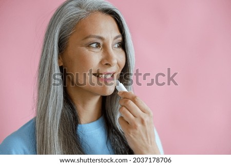 Positive silver haired Asian woman in blue t-shirt applies lip balm on pink background in studio, space for text. Mature beauty lifestyle