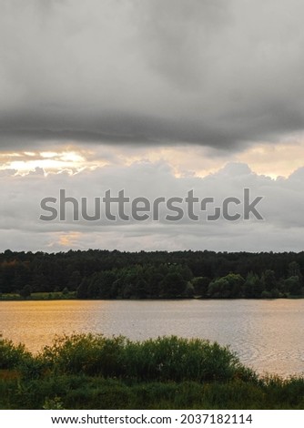 vertical photo, evening landscape, forest grows on the lake shore, large clouds are in the sky, the evening light of the sun is reflected in the water and clouds, Russia