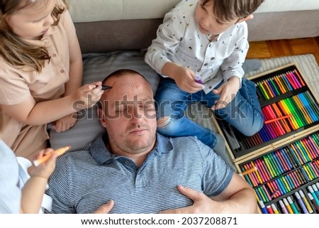 Adorable kids having fun with a sleeping father at home. Kids are having fun drawing on their father's face while he his sleeping on the floor. April fools prank. Lifestyle.