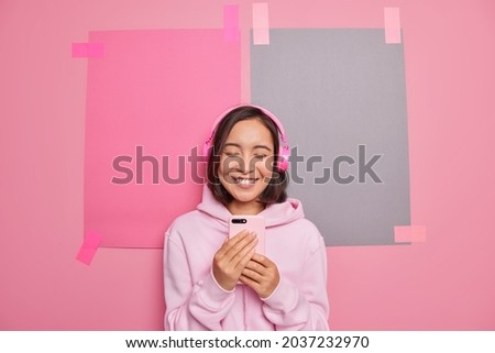 People free time modern technologies concept. Joyful Asian teenage girl uses mobile phone application for listening music wears stereo headphones on ears enjoys relaxing song after studying. Royalty-Free Stock Photo #2037232970