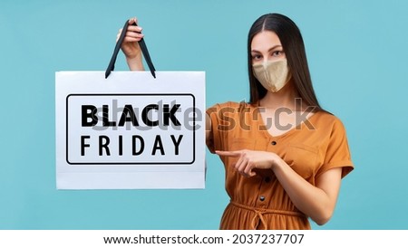 Studio portrait of confident young woman wearing orange dress and shiny protective mask, pointing at black friday shopping bag in her hand. Isolated on blue background. Mock up, copy space.           