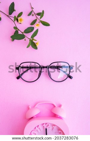 oval glasses in the photo in minimal summer style on a pastel pink color background. alarm clock, yellow flowers and green leaves
