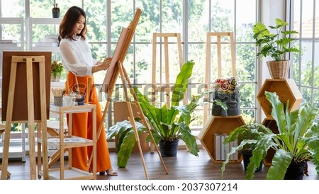 Senior woman artist painting image in studio at home with green small tree in little garden and full of natural light.