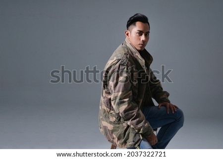 portrait of a male in camouflage coat ,jeans sitting chair on gray background
