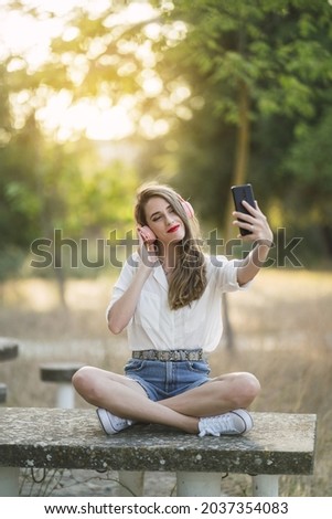 A young Spanish woman wearing shorts with a shirt and taking a selfie with a phone in a park