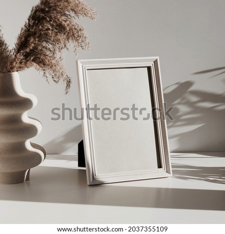 Frame mockup on white table, modern ceramic vase with dry grass and sunlight shadow.Neutral color. Photo frame, poster template on white wall background. Scandinavian interior. Copy space.