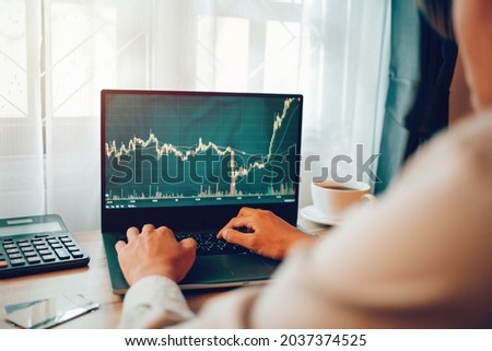 Happyness Asian young man 30s looking at stock tickers or graphs on cryptocurrency trading platforms on laptop with a cup of coffee, calculator, notebook on the table at his home office in the morning Royalty-Free Stock Photo #2037374525