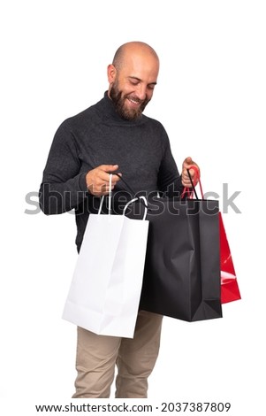 Smiling young caucasian man with beard looks inside shopping bag with satisfaction. Vertical image. Isolated on white background. Black friday sale concept. Birthday present concept.