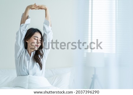 Asian beautiful girl in pajamas wake up in the morning with happiness. Attractive young woman smiling, feel happy and relax then stretching body after getting up from sleep on bed in bedroom at home. Royalty-Free Stock Photo #2037396290