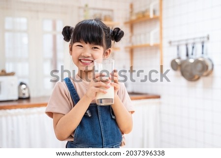 Portrait of Asian little cute kid holding a cup of milk in kitchen in house. Young preschool child girl or daughter stay home with smiling face, feel happy enjoy drinking milk and then look at camera. Royalty-Free Stock Photo #2037396338