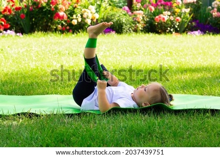 Little cute girl 1-3 in a white t-shirt goes in for sports with elastic on a green mat on the grass, against a background of flowers