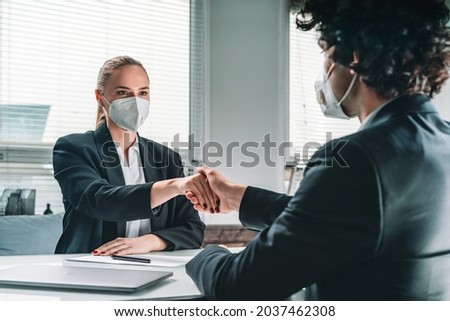 Businessman wearing formal suit is shaking hands with businesswoman in mask. Office workplace with laptop in the background. Concept of deal, agreement, teamwork, cooperation and communication
