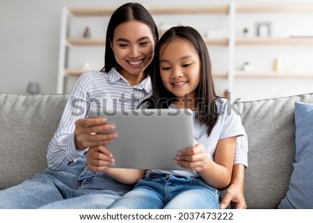 Happy asian mother and child girl using digital tablet together while sitting on couch at home, free space. Loving mom and daughter playing online games on pad or surfing internet Royalty-Free Stock Photo #2037473222