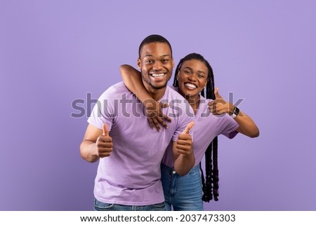 Best Choice, We Like It. Portrait of smiling black couple showing thumbs up gesture, hugging and approving or recommending something good. Man and woman posing isolated over purple studio background