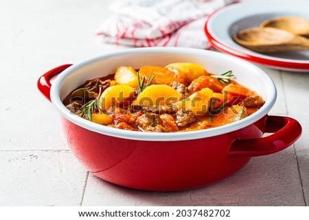 Beef stew with potatoes and carrots in tomato sauce in red pot, gray  background. Slow cooking concept. Royalty-Free Stock Photo #2037482702