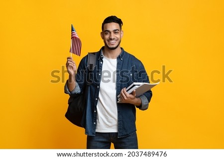 Smiling arab guy student with backpack and bunch of books showing flag of the US over yellow studio background, copy space. Positive middle-eastern young man studying English at school Royalty-Free Stock Photo #2037489476
