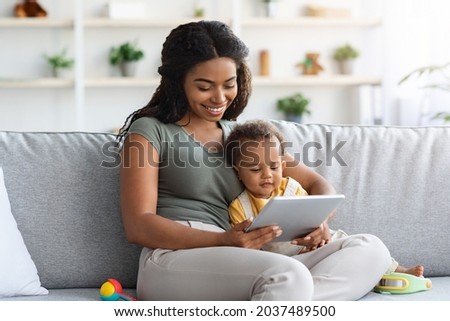 Happy Young Black Mother And Her Adorable Baby Son Relaxing With Digital Tablet At Home, Watching Development Videos Or Cartoons While Sitting Together On Couch In Living Room, Free Space