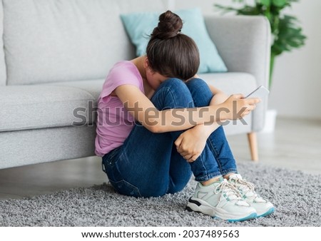 Depressed Indian teen girl with cellphone crying on floor at home, suffering from cyber bullying, being harassed online. Stressed teenager being victim of internet abuse or trolling Royalty-Free Stock Photo #2037489563