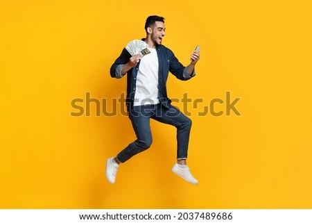 Happy handsome arabic guy with cash and mobile phone jumping up on yellow studio background, middle-eastern young man gambling or trading online, using mobile app, full length shot
