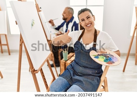 Young artist woman at art studio smiling happy and positive, thumb up doing excellent and approval sign 