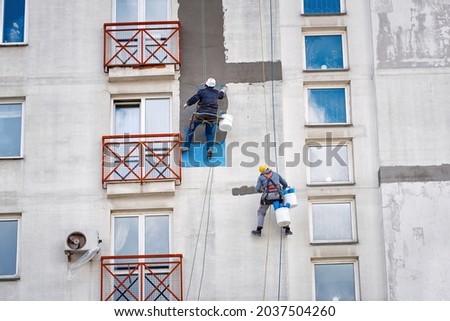  Industrial alpinist at height on rope, plastering wall with trowel. Industrial climber repairing house facade. Rope access job, construction workers repair and restore facade of high building. Royalty-Free Stock Photo #2037504260