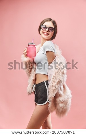 Smiling girl in faux pink fur coat black shorts white top and sunglasses holds pink beverage. Studio. Pink background. Red lips. Brunette.