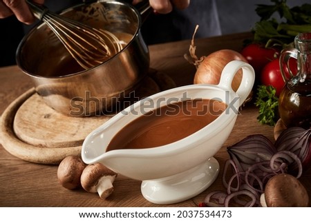 Sauce boat filled with rich brown spicy gravy viewed high angle on a kitchen table with assorted fresh vegetable ingredients Royalty-Free Stock Photo #2037534473