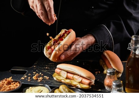 Chef preparing a tasty hot dog with a frankfurter on a fresh roll adding extra trimmings sprinkling crispy bacon bits onto the pickles and sausage Royalty-Free Stock Photo #2037534551