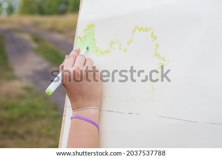 Woman's hand draws sketch of future drawing with green marker on white surface of paper  In background there is dirt road and foliage of trees 