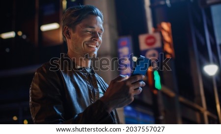 Portrait of Handsome Young Man Using Smartphone Standing in the Night City Street Full of Neon Lights. Smiling Stylish Blonde Male Using Mobile Phone for Social Media Posting. Royalty-Free Stock Photo #2037557027