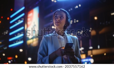 Beautiful Young Woman Using Smartphone Standing on the Night City Street Full of Neon Light. Portrait of Gorgeous Smiling Female Using Mobile Phone. Royalty-Free Stock Photo #2037557162