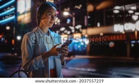 Beautiful Young Woman Using Smartphone Walking Through Night City Street Full of Neon Light. Portrait of Gorgeous Smiling Female Using Mobile Phone, Posting Social Media, Online Shopping, Texting